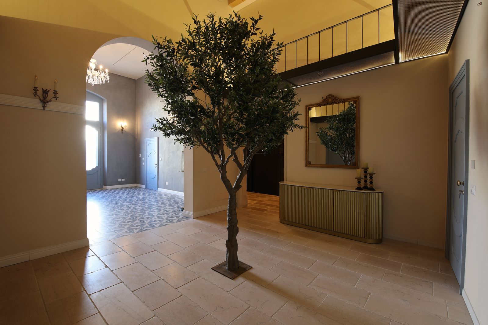 L'ingresso del palazzo, con pavimento in pietra, un ulivo al centro e una consolle dorata sotto uno specchio con cornice dorata.