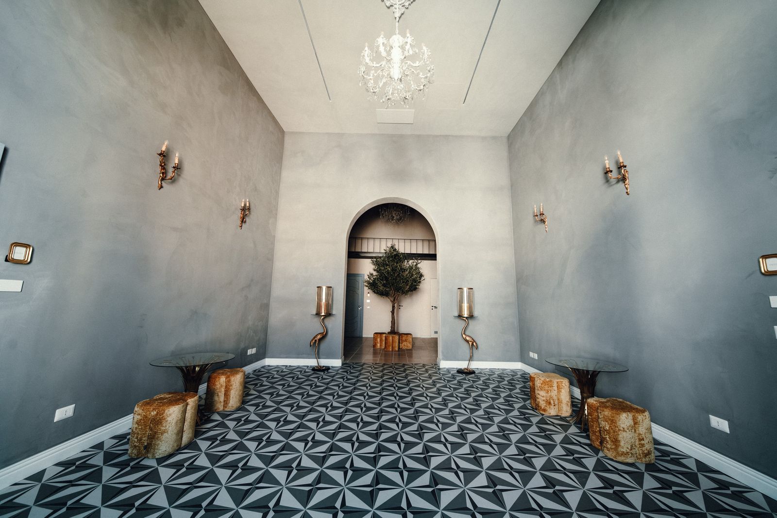 The long blue-grey hall with its geometric tiled floor, wall sconces and tree-stump stools, leading to an arched doorway and an olive tree.