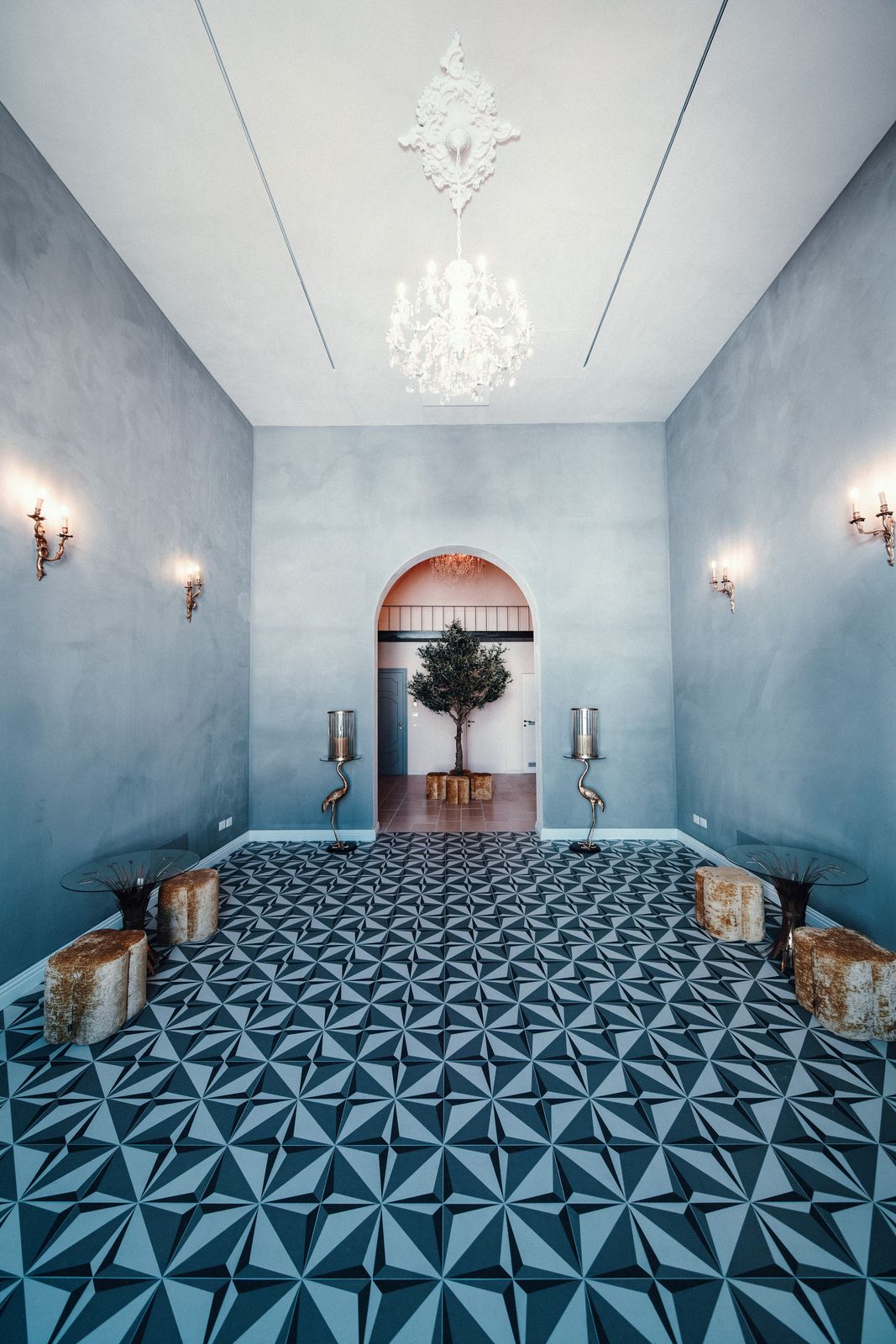 A crystal chandelier hanging high above the blue-grey hall and its geometric floor, with the arched doorway at the far end.