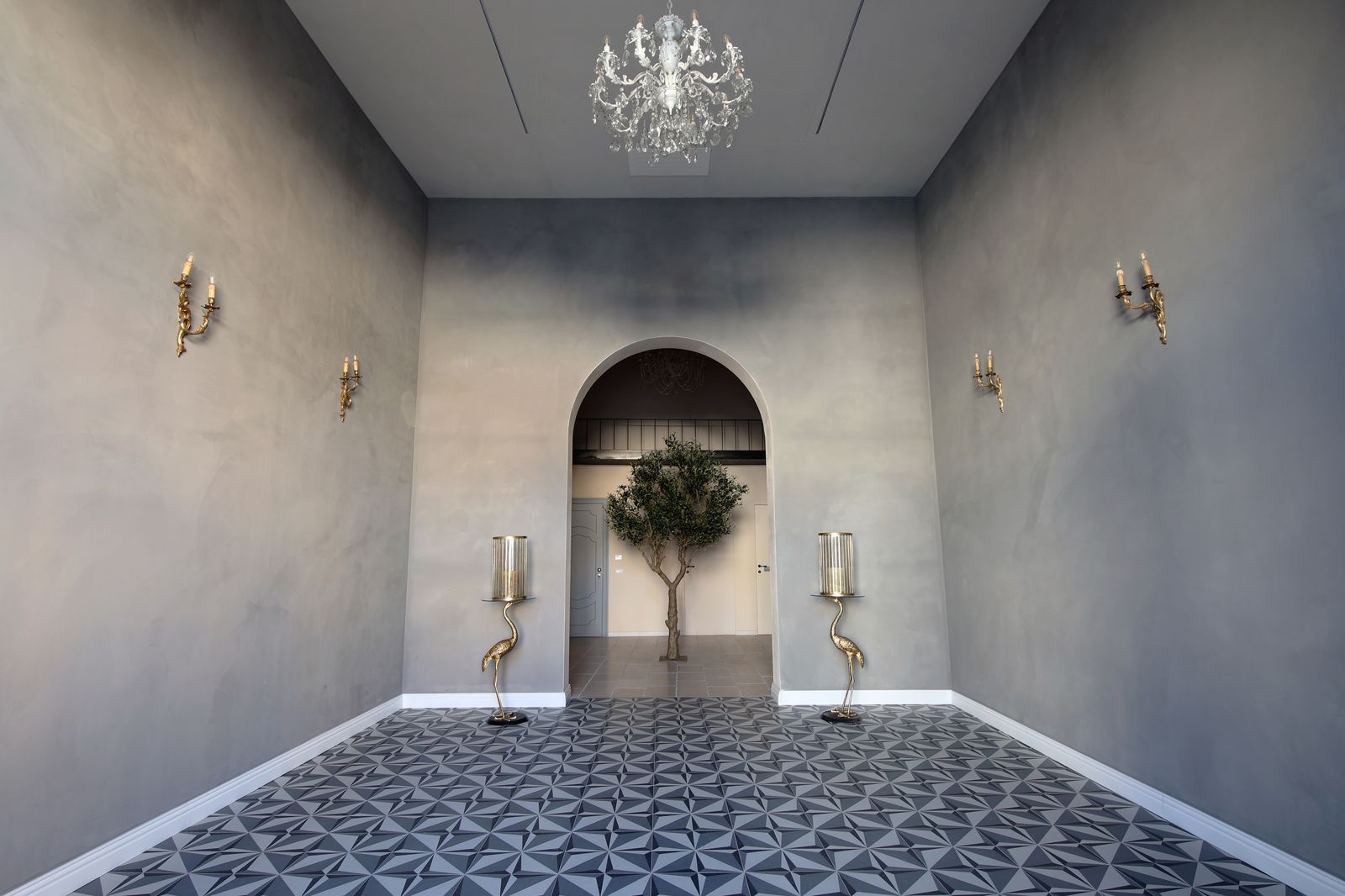 Sala dalle pareti grigie con lampadario di cristallo, candelabri in ottone a forma di gru e pavimento a motivi geometrici, aperta ad arco verso l'ulivo.