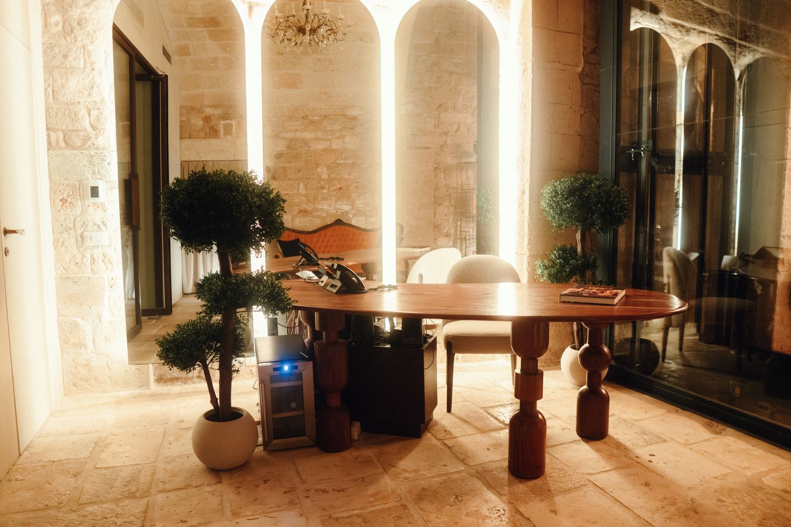 The entrance hall at night, with a round wooden table, clipped topiary in white pots and softly lit stone arches behind.