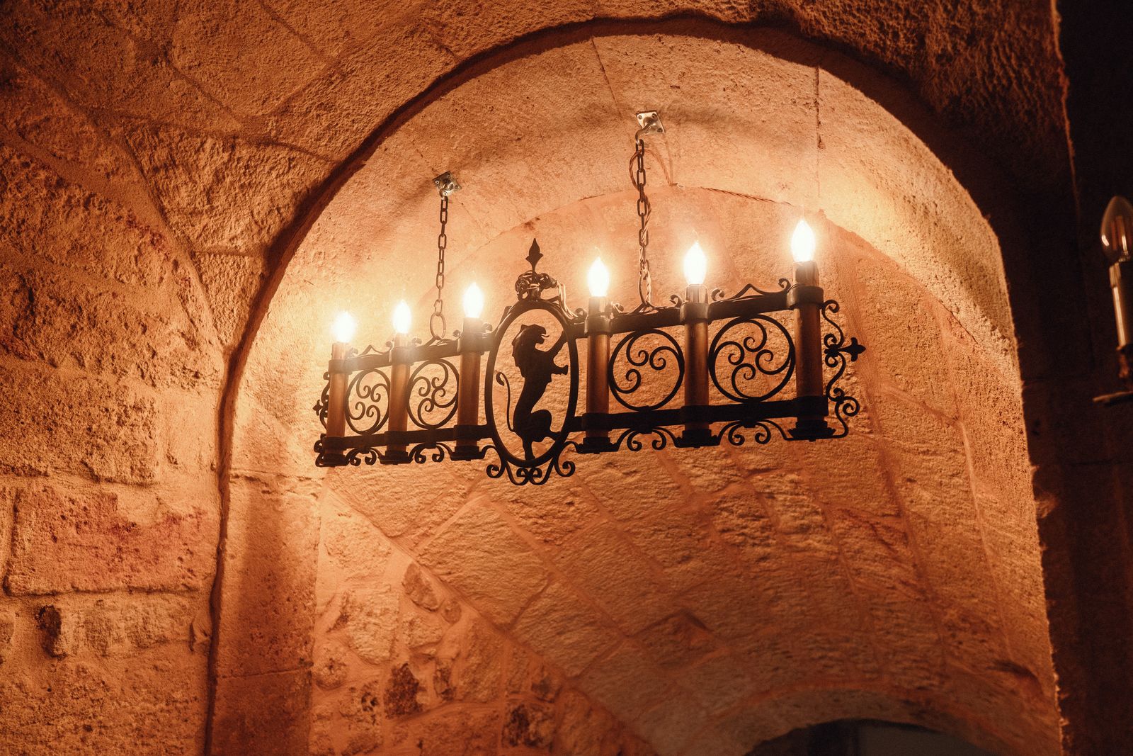 A wrought-iron candle chandelier with a heraldic emblem, hanging beneath a stone vault.