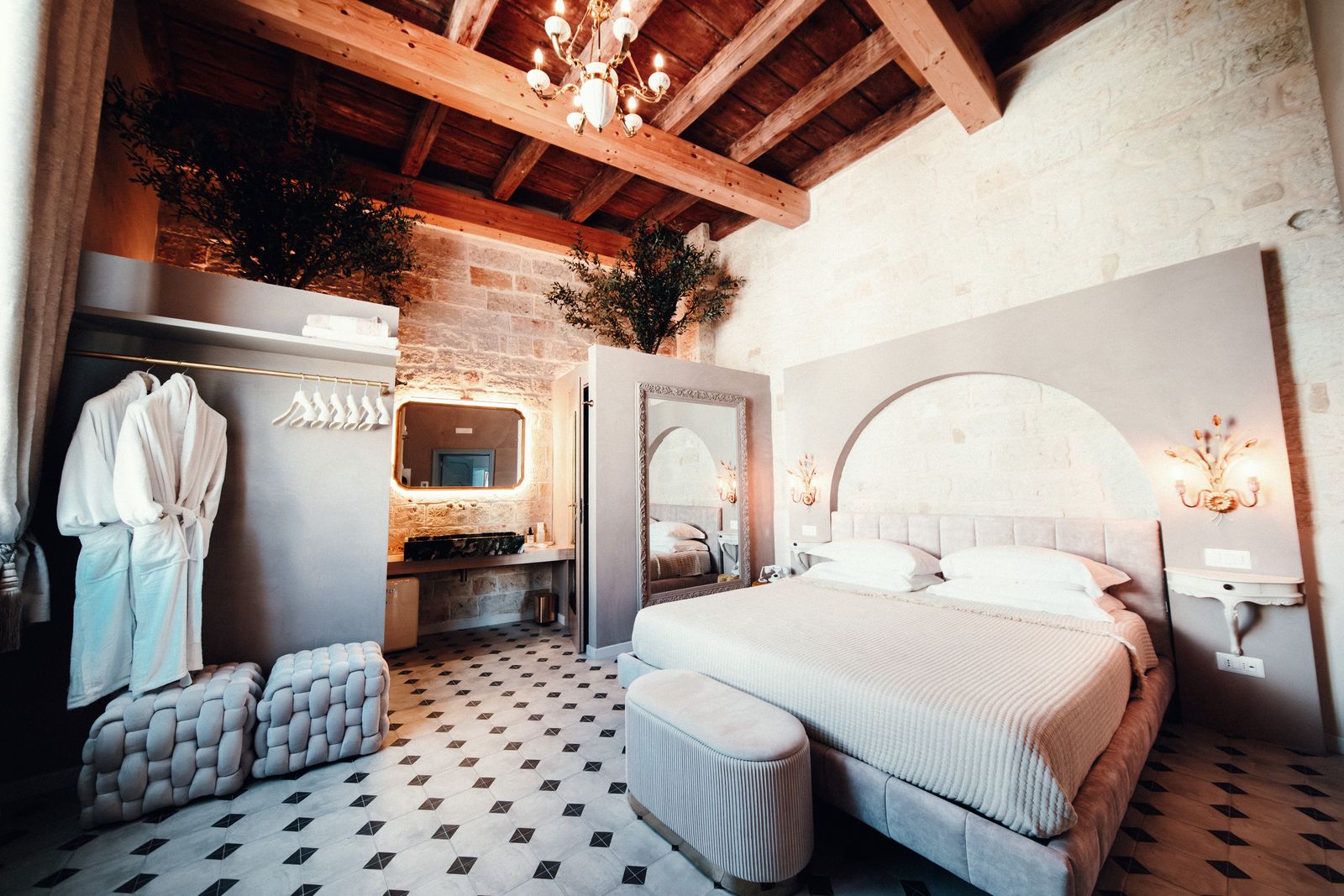Garden Deluxe Room: a bed with an arched headboard beneath the beamed ceiling and a crystal chandelier, with olive trees, white robes and woven poufs by the vanity.