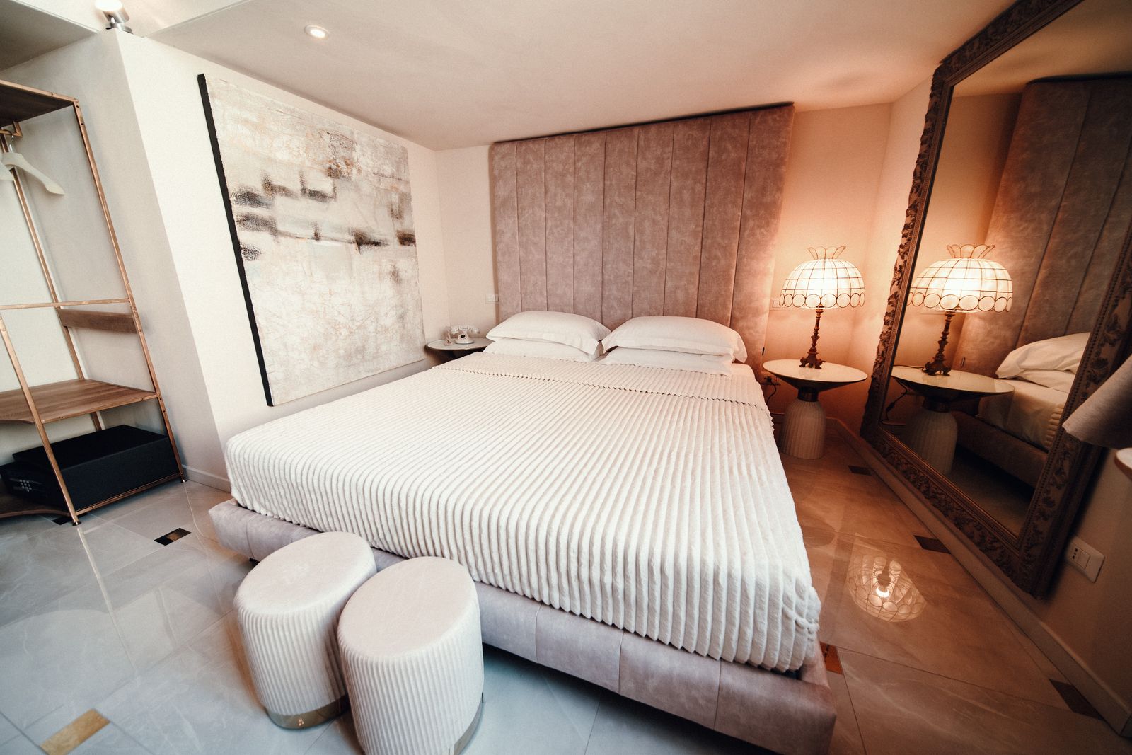 Reflection Junior Suite: a bed with a tall padded headboard and two round poufs at its foot, beside an ornate floor mirror reflecting the bedside lamps.