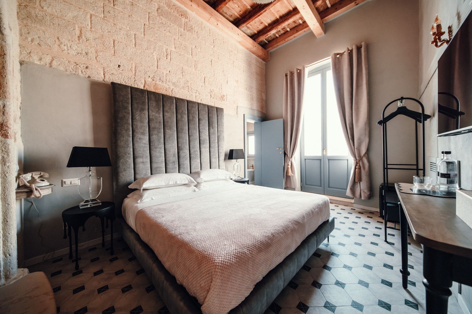 Venus Deluxe Room: a bed with a tall velvet headboard against the stone wall, beneath a beamed ceiling, with French windows and soft curtains beyond.