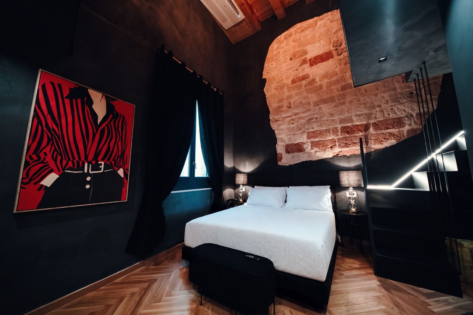 Firestone Junior Suite: dark walls and herringbone parquet, with a bold red portrait painting, a lit patch of raw stone above the bed and a black staircase to the upper level.