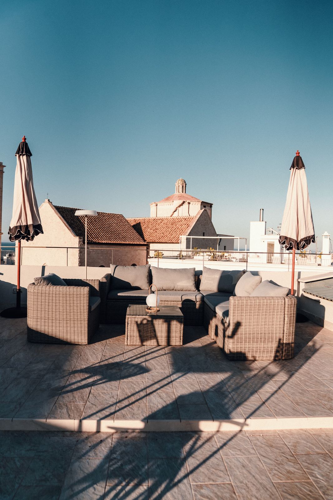 Wicker sofas and armchairs on the rooftop terrace between two parasols, with a church dome against a clear blue sky.